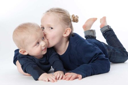 geschwisterportrait im studio, klassisch vor weiß, große schwester küsst kleinen bruder auf die wange