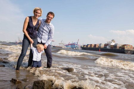 eltern mit baby am elbstrand im wasser mit containerschiff im hintergrund