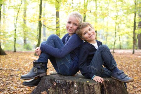 junge und mädchen sitzen im wald auf einem baumstamm und grinsen in die kamera