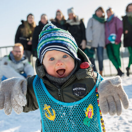Kleinkind im Schneeanzug mit viel zu großen Handschuhen läuft von einer Gruppe weg auf die Kamera zu