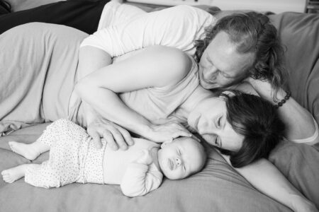 portrait von eltern, sie liegen mit schalfendem baby auf der couch und schauen ganz liebevoll