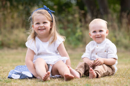 Geschwisterportrait von zwei Kleinkindern, einem Mädchen, einem Jungen, beide lachen in die Kamera und sitzen im park