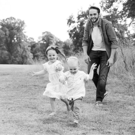 familienfoto eines lachenden Vaters, der seine zwei lachenden kleinkinder über die wiese jagt